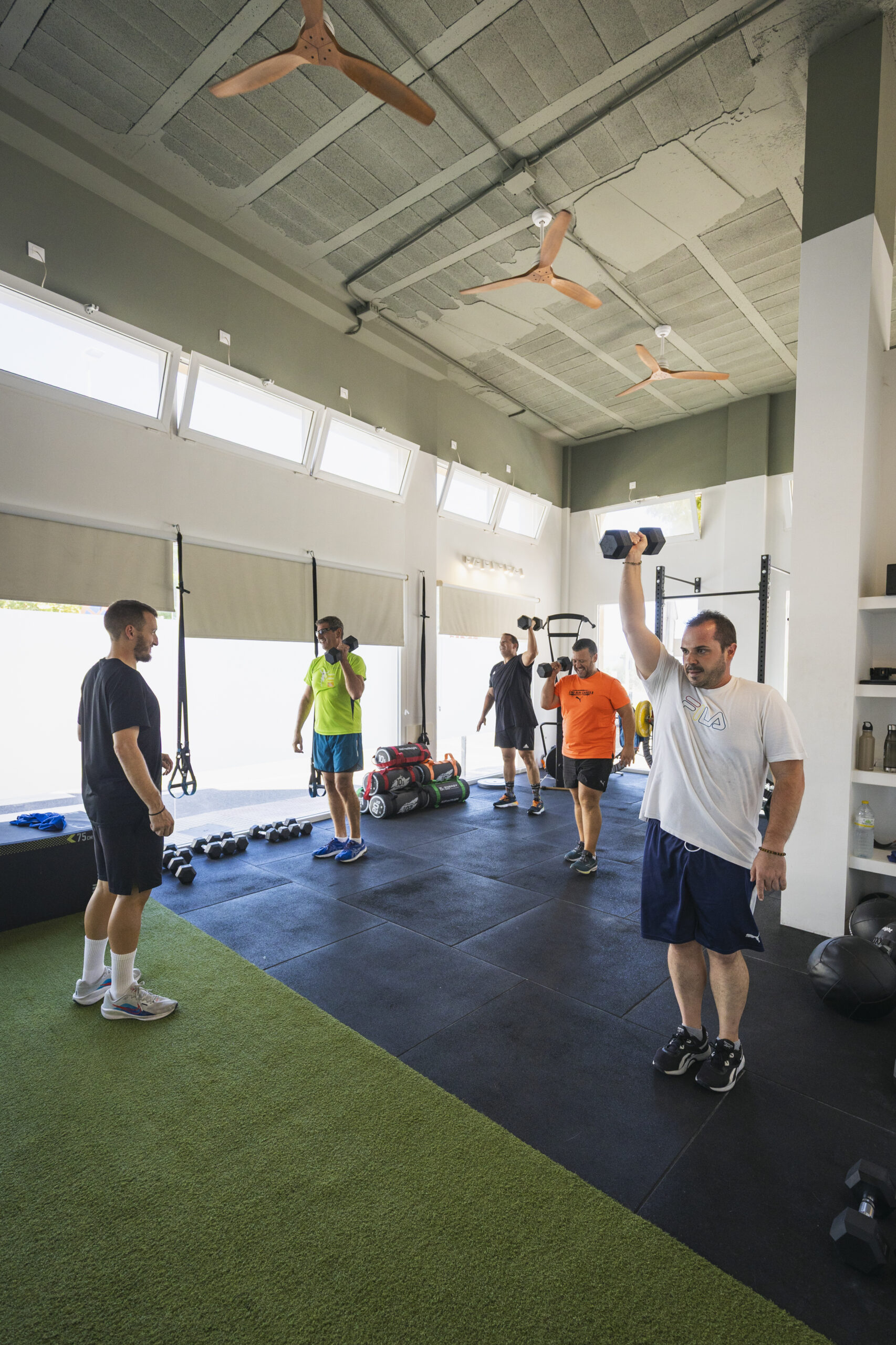 Grupo de personas realizando entrenamiento de fuerza con mancuernas en un gimnasio. Varios individuos levantan pesas en un espacio abierto con suelo deportivo.