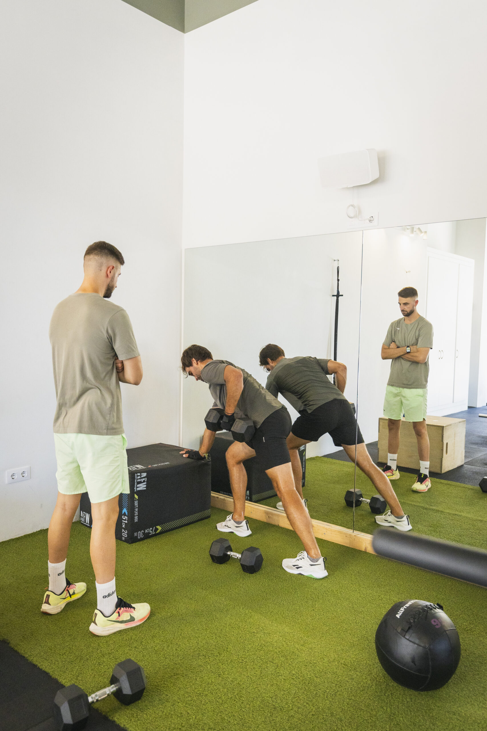 Entrenamiento de fuerza y resistencia en un gimnasio. Persona realizando un ejercicio de remo con mancuerna apoyándose en un cajón, bajo la supervisión de un entrenador personal en Jávea