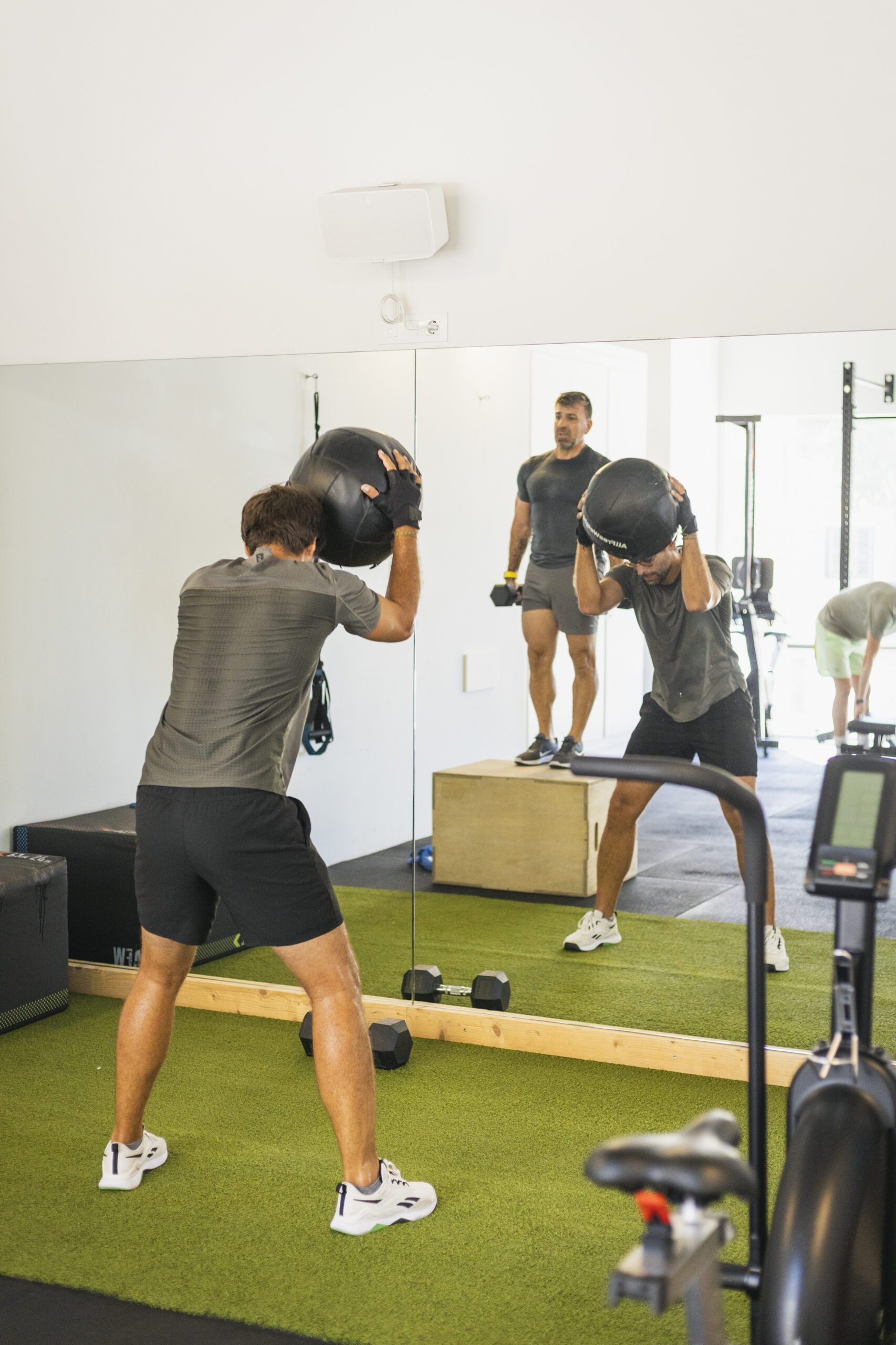 Entrenamiento funcional con balón medicinal y box jump en un gimnasio o centro de entrenamiento personal. Persona levantando un peso sobre la cabeza para ejercicios de core y fuerza explosiva, con la ayuda de un entrenador en el reflejo.