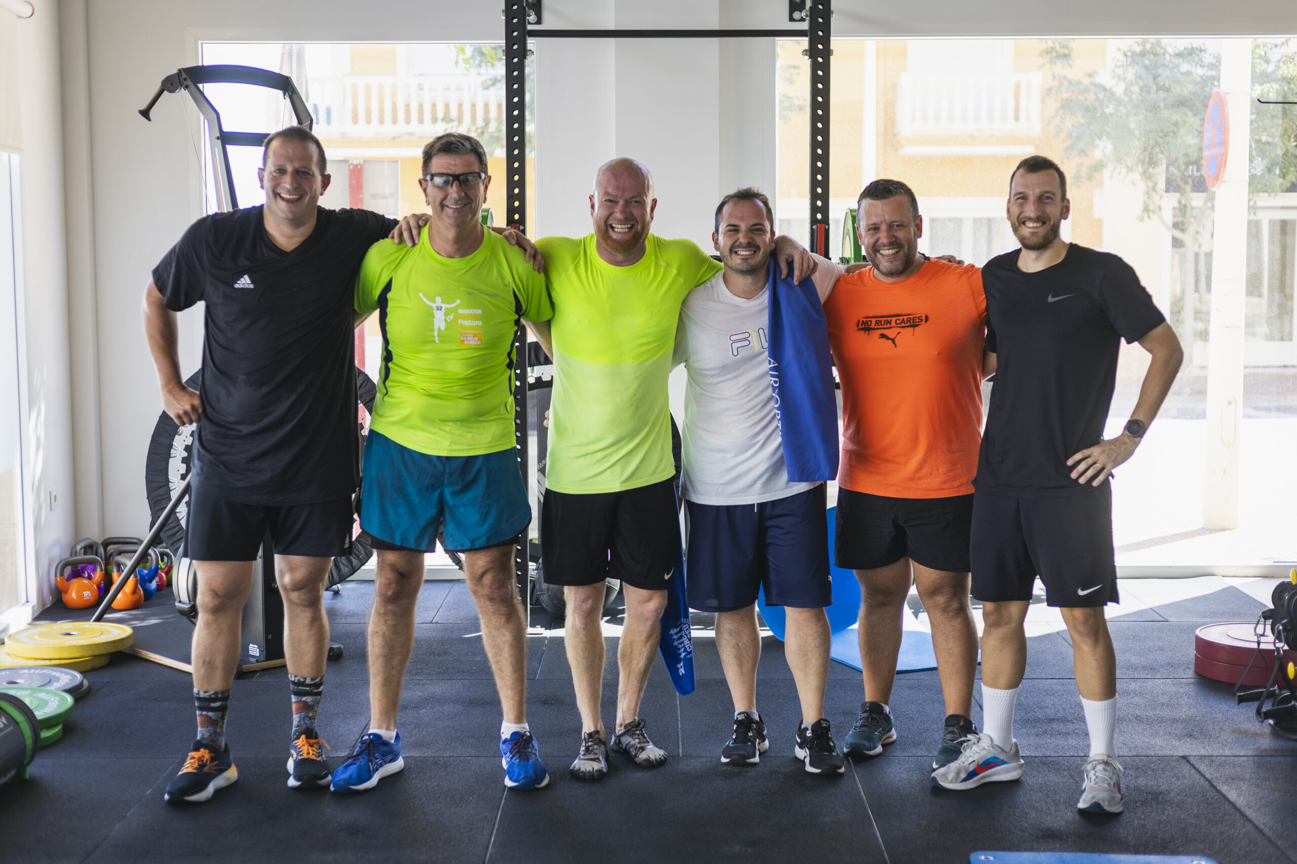 Grupo de seis hombres sonriendo y posando con sus brazos alrededor, celebrando la finalización de una sesión de fitness o entrenamiento en un gimnasio. Refleja comunidad, deporte y bienestar.